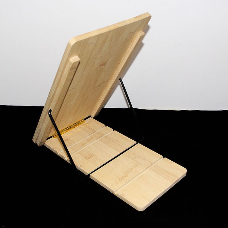May include: A light-coloured bamboo book stand with a hinged design. The stand features a sloped reading surface supported by a black metal frame. The stand is designed to hold books or tablets.
