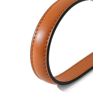 May include: Close-up of a curved, brown leather strap with black edging and visible stitching. The strap appears to be part of a bag or handle, showcasing a smooth, textured surface.