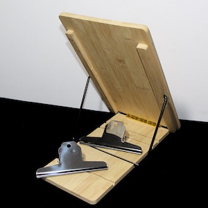May include: A bamboo cookbook stand with a slanted design. It features a rectangular base and a back panel supported by black metal rods. Two silver metal clips are attached to the base to hold pages in place.