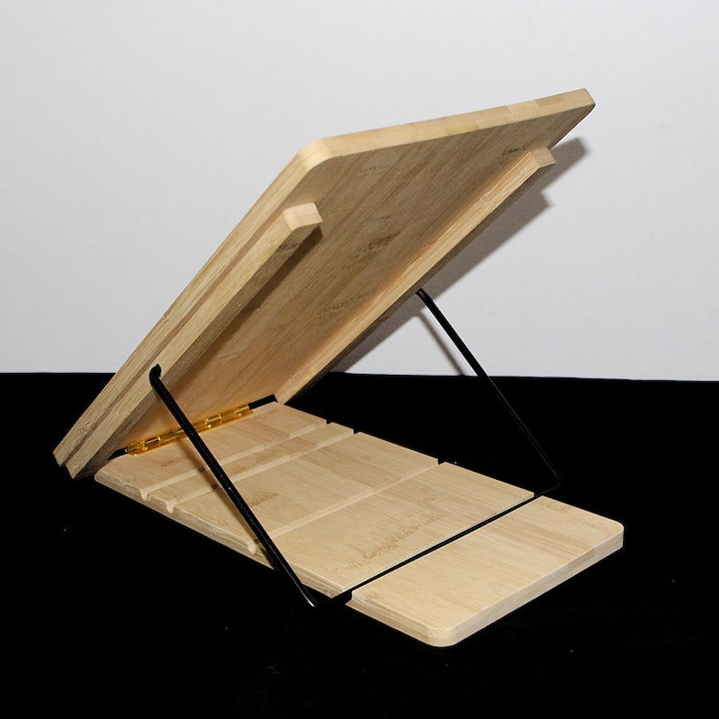 May include: A light-coloured bamboo book stand with a slatted base and an angled top. The stand is supported by black metal legs and features a brass-coloured hinge. The stand is designed to hold books or tablets.