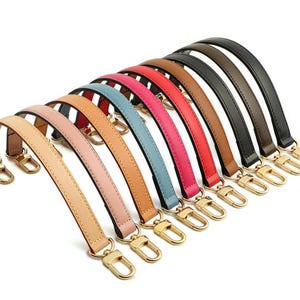 May include: A collection of colorful leather bag handles in various shades, including tan, pink, blue, red, brown, and black. Each handle is curved and features gold-colored metal clasps for attachment.