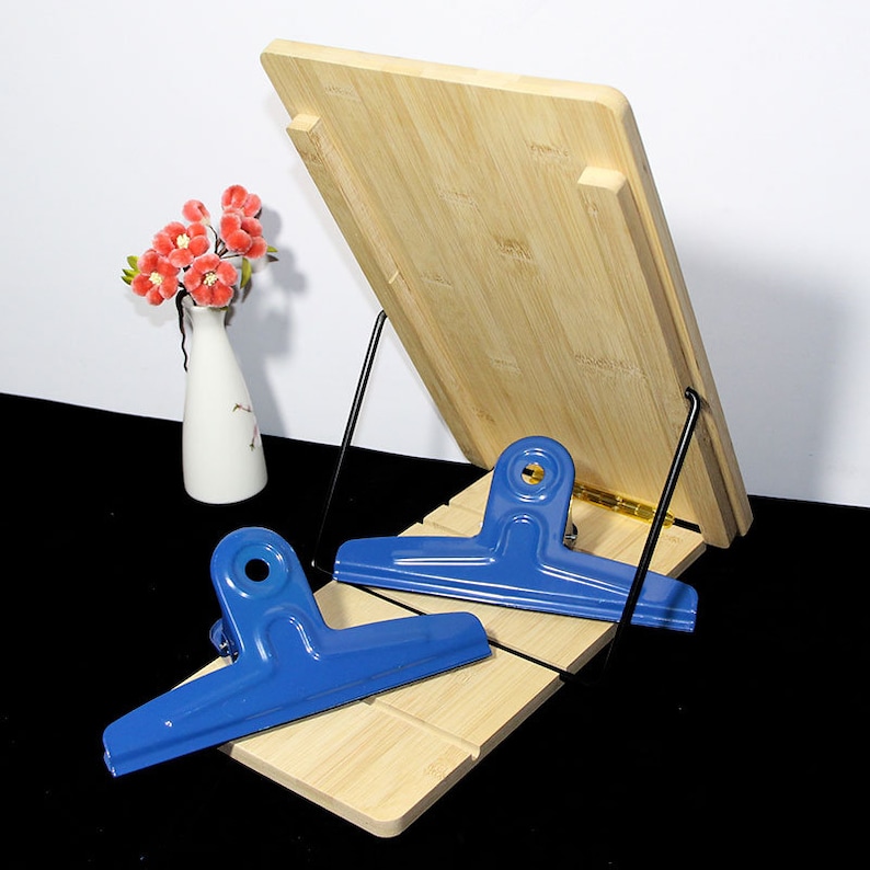 May include: A bamboo cookbook stand with a blue metal clip. The stand is angled with a black metal support. A small white vase with red flowers is in the background. The stand is 30 cm wide and 23 cm tall.