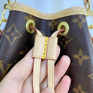 May include: Close-up of the drawstring detail on a brown and tan Louis Vuitton bucket bag. The bag has a brown monogram canvas with tan leather trim and gold hardware.