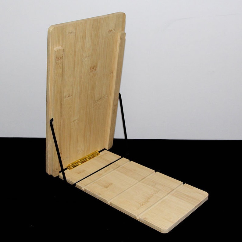 May include: A bamboo book stand with a natural wood finish. The stand has a hinged design with a back support and a slatted base. The stand is supported by black metal legs and is designed to hold books or tablets.