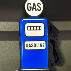 May include: A blue and white vintage-style petrol pump sign with the word "GAS" in a white circle at the top. The word "GASOLINE" is displayed on the front. The sign has a black border and a white cord.
