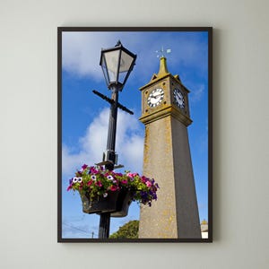 May include: A black street lamp with a flower box filled with pink, purple, and white flowers is in front of a tall, grey stone clock tower with a clock face.
