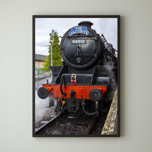 May include: Framed print of a black steam train, "The Jacobite," with the number 44871 on the front. The train has a red and orange front, and is emitting steam. The image is framed in a dark brown frame.