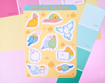 May include: A set of colorful stickers featuring budgies in various poses, including a budgie wearing a bell, a budgie on a laptop, and a budgie in a cage. The stickers are on a yellow background with a white border and the text "BUDGIE BUDDIES" at the top.