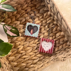 May include: Two square keychains with scalloped edges and metal rings. One features a light blue and white gingham pattern with a heart-shaped photo insert. The other has a red and white striped pattern with a heart-shaped insert reading Your cute face here!