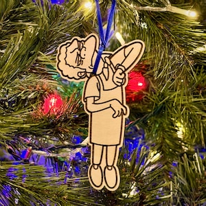 May include: Wooden Christmas tree ornament featuring a cartoon character in a pose. The ornament is made of light-colored wood with black outlines and a blue ribbon for hanging. The ornament is hanging on a Christmas tree with colorful lights.