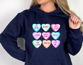 May include: A navy blue hooded sweatshirt featuring a graphic print of nine pastel-colored conversation hearts. Each heart contains red text including BE MINE, XOXO, HOT STUFF, LOVE, SWEET HEART, CUTIE, FOR EVER, DARLIN, and TXT ME in a grid pattern.