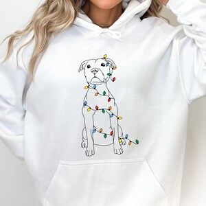 May include: White hoodie featuring a line drawing of a dog adorned with colorful Christmas lights. The dog is sitting, and the lights are wrapped around its body. The hoodie has a front pocket and a drawstring hood.