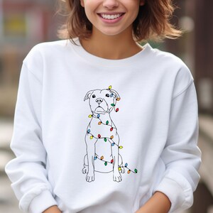 May include: White sweatshirt featuring a line drawing of a dog adorned with colorful Christmas lights. The dog is sitting upright, and the lights are wrapped around its body. The sweatshirt is a solid white color.