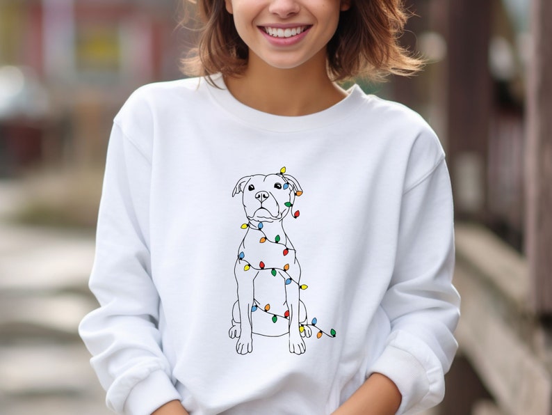 May include: White sweatshirt featuring a line drawing of a dog adorned with colorful Christmas lights. The dog is sitting upright, and the lights are wrapped around its body. The sweatshirt is a solid white color.