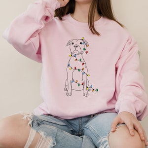 May include: A light pink sweatshirt featuring a black line drawing of a dog with colorful Christmas lights draped around it. The dog is sitting upright. The sweatshirt has long sleeves and a crew neck.