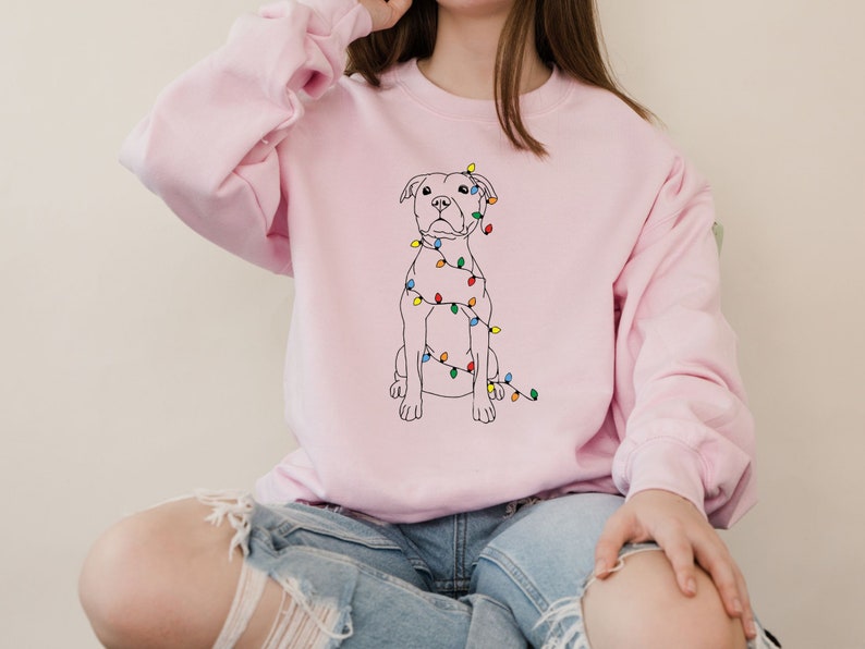 May include: A light pink sweatshirt featuring a black line drawing of a dog with colorful Christmas lights draped around it. The dog is sitting upright. The sweatshirt has long sleeves and a crew neck.