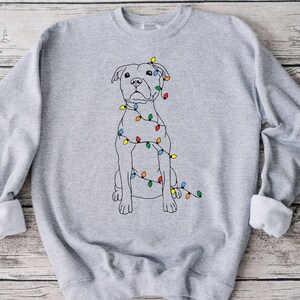May include: A light gray sweatshirt featuring a line drawing of a dog wrapped in colorful Christmas lights. The dog is sitting upright with a neutral expression. The sweatshirt has a crew neck and long sleeves with rolled cuffs.
