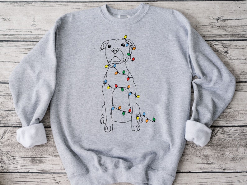 May include: A light gray sweatshirt featuring a line drawing of a dog wrapped in colorful Christmas lights. The dog is sitting upright with a neutral expression. The sweatshirt has a crew neck and long sleeves with rolled cuffs.