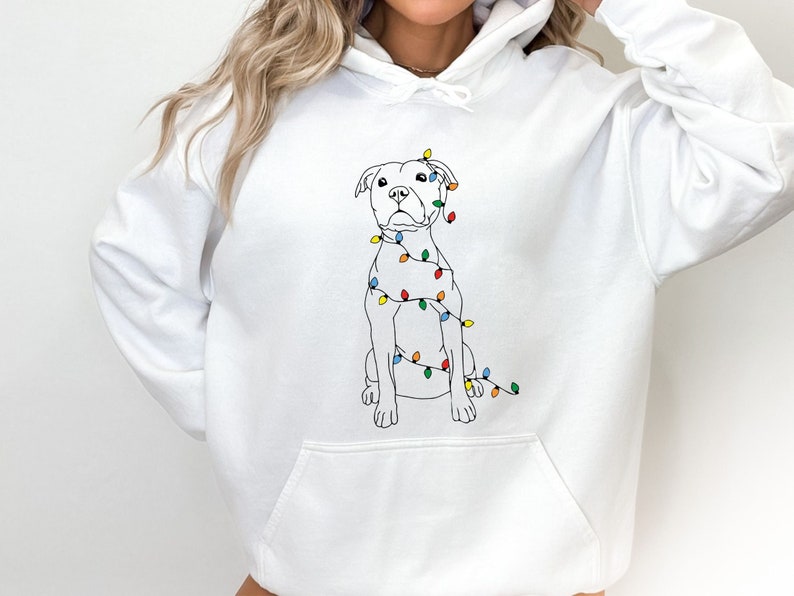 May include: White hoodie featuring a line drawing of a dog adorned with colorful Christmas lights. The dog is sitting, and the lights are wrapped around its body. The hoodie has a front pocket and a drawstring hood.
