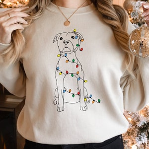 May include: A cream-colored sweatshirt featuring a line drawing of a dog wrapped in colorful Christmas lights. The dog is sitting upright, with the lights draped around its body. The design is simple and festive, perfect for the holiday season.