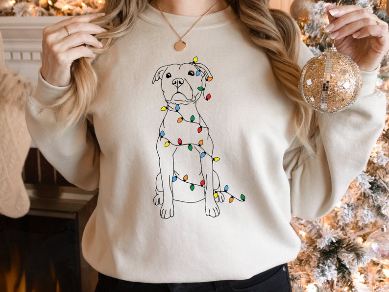 May include: A cream-colored sweatshirt featuring a line drawing of a dog wrapped in colorful Christmas lights. The dog is sitting upright, with the lights draped around its body. The design is simple and festive, perfect for the holiday season.