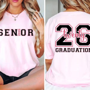 May include: Light pink t-shirt with black and pink text. The front of the shirt says "SENIOR 2026". The back of the shirt has a large "26" with "Twenty" in the middle and "GRADUATION" below.