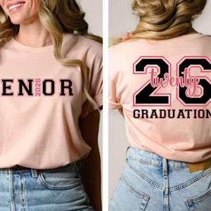 May include: A light pink t-shirt with the word "SENIOR 2026" printed on the front in black and pink lettering. The back of the shirt features the number "26" with the word "Twenty" inside and the word "GRADUATION" below.