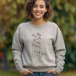 May include: A light gray sweatshirt featuring a line drawing of a dog wrapped in colorful Christmas lights. The dog is sitting upright. The sweatshirt is a crew neck style.