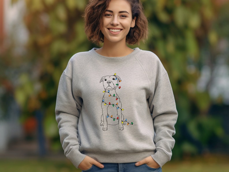 May include: A light gray sweatshirt featuring a line drawing of a dog wrapped in colorful Christmas lights. The dog is sitting upright. The sweatshirt is a crew neck style.