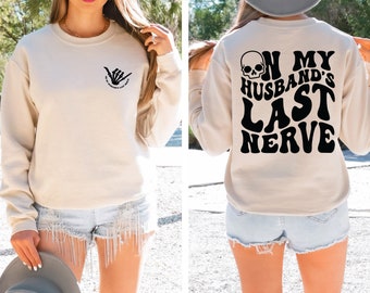 May include: A cream colored sweatshirt with a black skull and the text "On My Husband's Last Nerve" printed on the back. The front of the sweatshirt has a small black skeleton hand making a shaka sign with the text "On My Husband's Last Nerve" underneath.