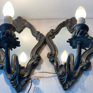 May include: Pair of ornate, antique-style wall sconces with mirrored backs. Each sconce features a curved arm holding a light bulb, with decorative floral and scrollwork details. The sconces are made of a dark metal and are mounted on a white surface.