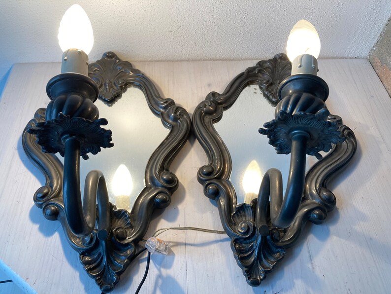 May include: Pair of ornate, antique-style wall sconces with mirrored backs. Each sconce features a curved arm holding a light bulb, with decorative floral and scrollwork details. The sconces are made of a dark metal and are mounted on a white surface.