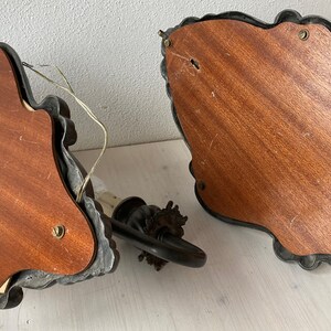 May include: Two antique wall sconces with ornate, dark metal frames and wooden backing. The sconces have a decorative, leaf-like design around the edges. One sconce has a light fixture attached, with a black arm and a bulb socket.