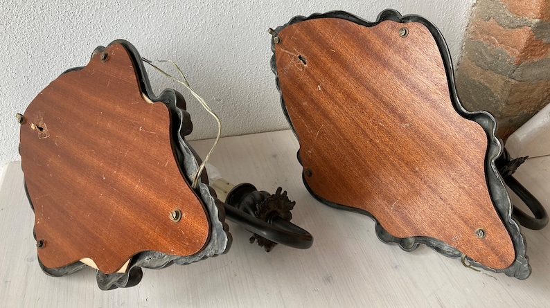 May include: Two antique wall sconces with ornate, dark metal frames and wooden backing. The sconces have a decorative, leaf-like design around the edges. One sconce has a light fixture attached, with a black arm and a bulb socket.