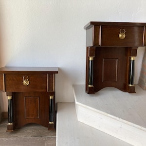 以下が含まれることがあります： 引き出しと真鍮製の金具が付いた、お揃いの濃い色の木製ナイトスタンド2つ。各ナイトスタンドには、黒と金のアクセントの柱があります。ナイトスタンドは白い表面とタイルの床に展示されています。