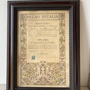 May include: Framed Italian diploma from the Ministry of Education. The document features ornate borders and text in Italian, with a stamp and decorative elements in green, beige, and brown. The dark brown frame adds a classic touch.