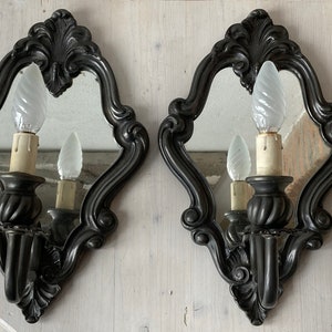 May include: Pair of ornate, dark gray wall sconces with mirrors and decorative frames. Each sconce features a candle-style light bulb. The sconces are designed with a classic, vintage aesthetic, suitable for home decor.