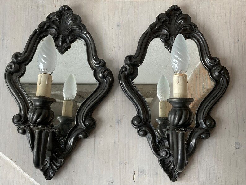 May include: Pair of ornate, dark gray wall sconces with mirrors and decorative frames. Each sconce features a candle-style light bulb. The sconces are designed with a classic, vintage aesthetic, suitable for home decor.