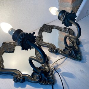 May include: Two antique wall sconces with ornate, dark metal frames and integrated mirrors. Each sconce features a curved arm holding a light bulb. The frames have a decorative, floral design. The sconces are wired for electricity.