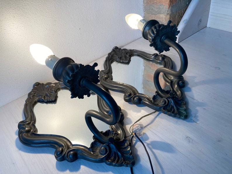 May include: Two antique wall sconces with ornate, dark metal frames and integrated mirrors. Each sconce features a curved arm holding a light bulb. The frames have a decorative, floral design. The sconces are wired for electricity.
