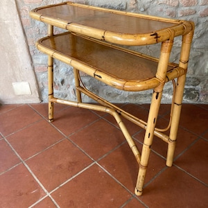Puede incluir: Una consola de bambú de dos niveles con acabado natural. La mesa tiene una parte superior rectangular y un estante inferior, ambos con una superficie marrón claro. El marco está hecho de postes de bambú, unidos con bambú tejido. La mesa mide aproximadamente 76 cm de alto.