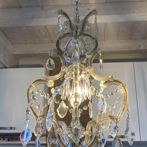 May include: A crystal chandelier with a gold finish and clear glass crystals. The chandelier is hanging from a white ceiling and has a traditional design.