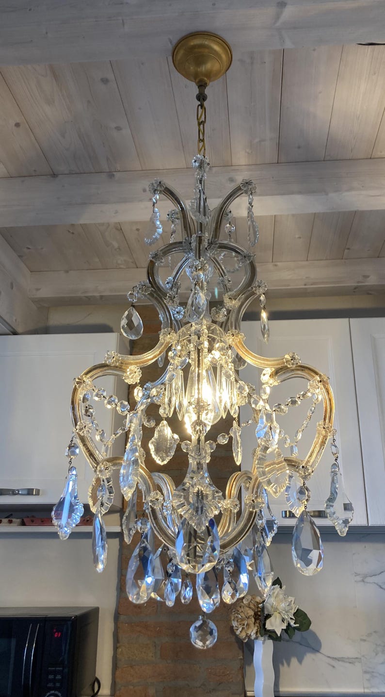 May include: A crystal chandelier with a gold finish and clear glass crystals. The chandelier is hanging from a white ceiling and has a traditional design.