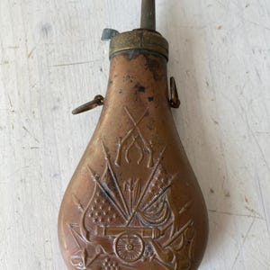 Peut inclure: Une poire à poudre en cuivre avec un motif en relief représentant des fusils croisés, des drapeaux et un canon. L'objet possède un embout en laiton avec un bec étroit et un petit levier métallique. Deux anneaux sont fixés sur les côtés.