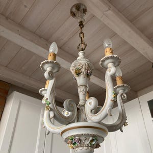 May include: A white and gold chandelier with three curved arms, each holding a light bulb. The chandelier is decorated with floral accents in various colors. The fixture hangs from a chain attached to the ceiling.