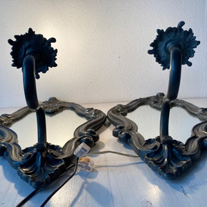 May include: Pair of ornate wall sconces with mirrored surfaces and dark metal frames. Each sconce features a curved arm supporting a decorative top and a mirrored base with an intricate border. The sconces are designed for wall mounting.