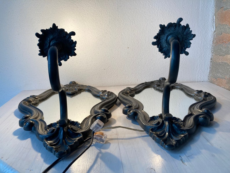 May include: Pair of ornate wall sconces with mirrored surfaces and dark metal frames. Each sconce features a curved arm supporting a decorative top and a mirrored base with an intricate border. The sconces are designed for wall mounting.