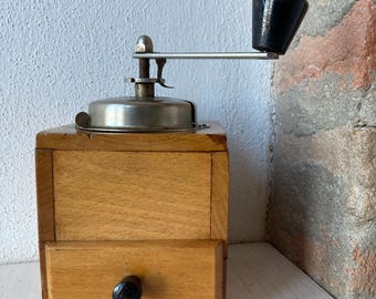 May include: A vintage wooden coffee grinder with a metal crank and a drawer. The grinder is made of light wood and has a dark brown handle.