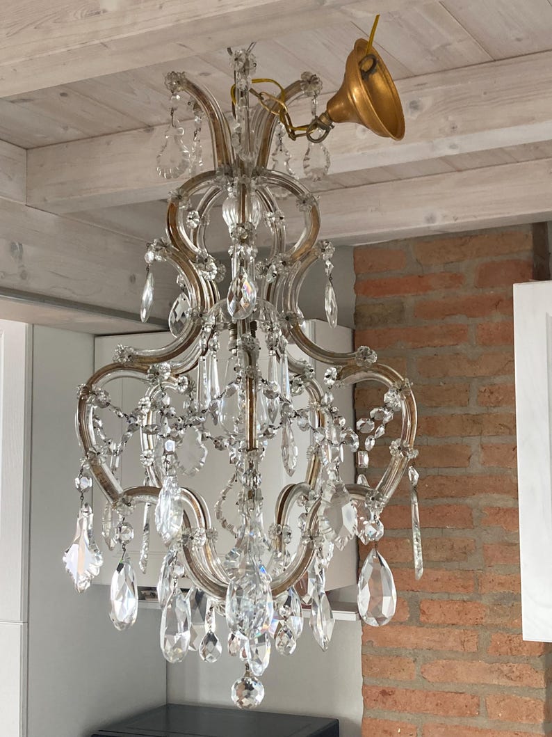 May include: A crystal chandelier with a gold-toned metal frame and many clear crystal drops. The chandelier is hanging from a white ceiling.
