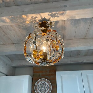 May include: A gold-toned chandelier with clear and amber glass flower accents hangs from a white wood ceiling. The chandelier is illuminated by a single light bulb.